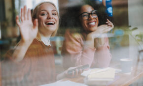 Two young smiling women seen through a window; one is waving.