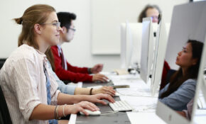 A student works on a computer in a classroom. Another student, also working, is reflected in her monitor.